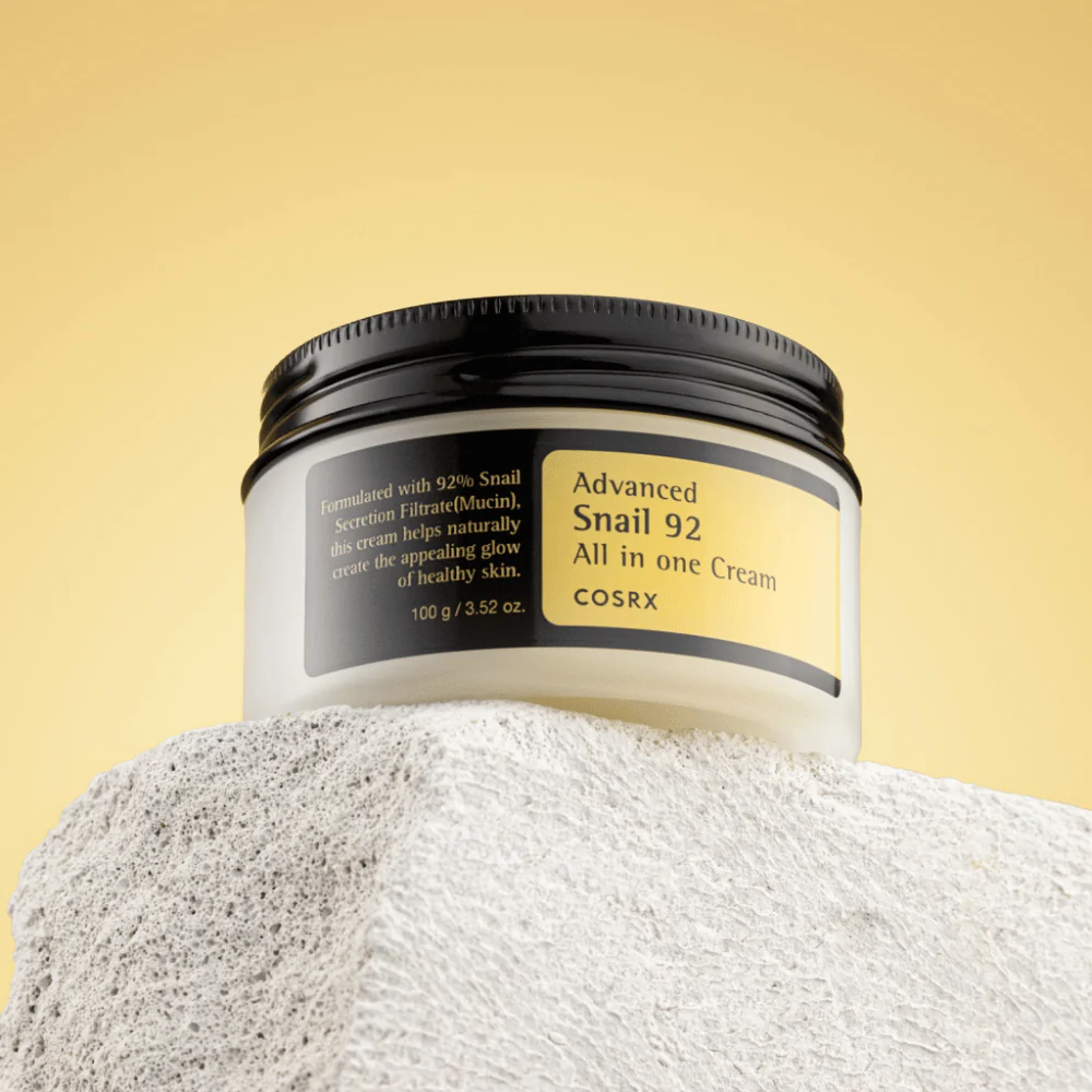 COSRX Advanced Snail 92 All in One Cream – হাল্কা জেল-ক্রিমি টেক্সচারের ৯২% স্নেইল মিউসিন সমৃদ্ধ ময়েশ্চারাইজার, এতে কালো ঢাকনা ও স্বচ্ছ কাচের জারের ভিতরের দিকে হালকা হলুদ লেবেল রয়েছে।"